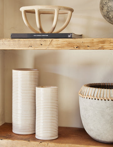 #color::sand #size::large | The Prong cream stoneware ceramic centerpiece bowl by SIN sits on a wooden shelf with narrow ribbed vases, a black paperback book, and a vase with jute detailing