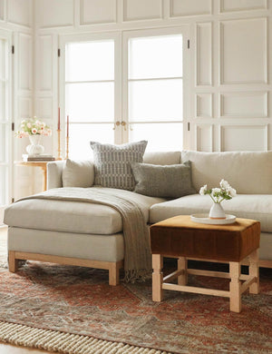  The Ambleside cognac velvet ottoman sits atop a red Persian-inspired rug in front of a linen sectional sofa
