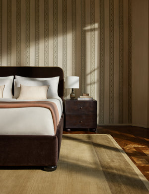 A velvet upholstered platform bed and nightstand sit infront of a wall featuring the Paradiso striped Wallpaper by Jake Arnold