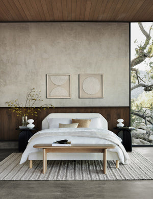 The Henrik light wood bench sits at the end of a white linen framed bed atop a textured ivory carpet in a room with concrete floors