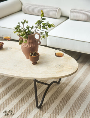 The Peggy sculptural iron frame and stone top oval outdoor coffee table styled with a modern white outdoor sofa and striped outdoor rug.