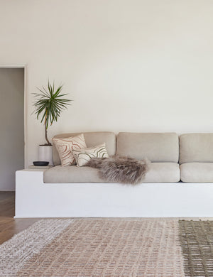 The Terra handcrafted textured multicolored floor rug by Élan Byrd sits in a living room with a gray linen cushioned bench, a gray sheepskin throw, and green and terracotta throw pillows