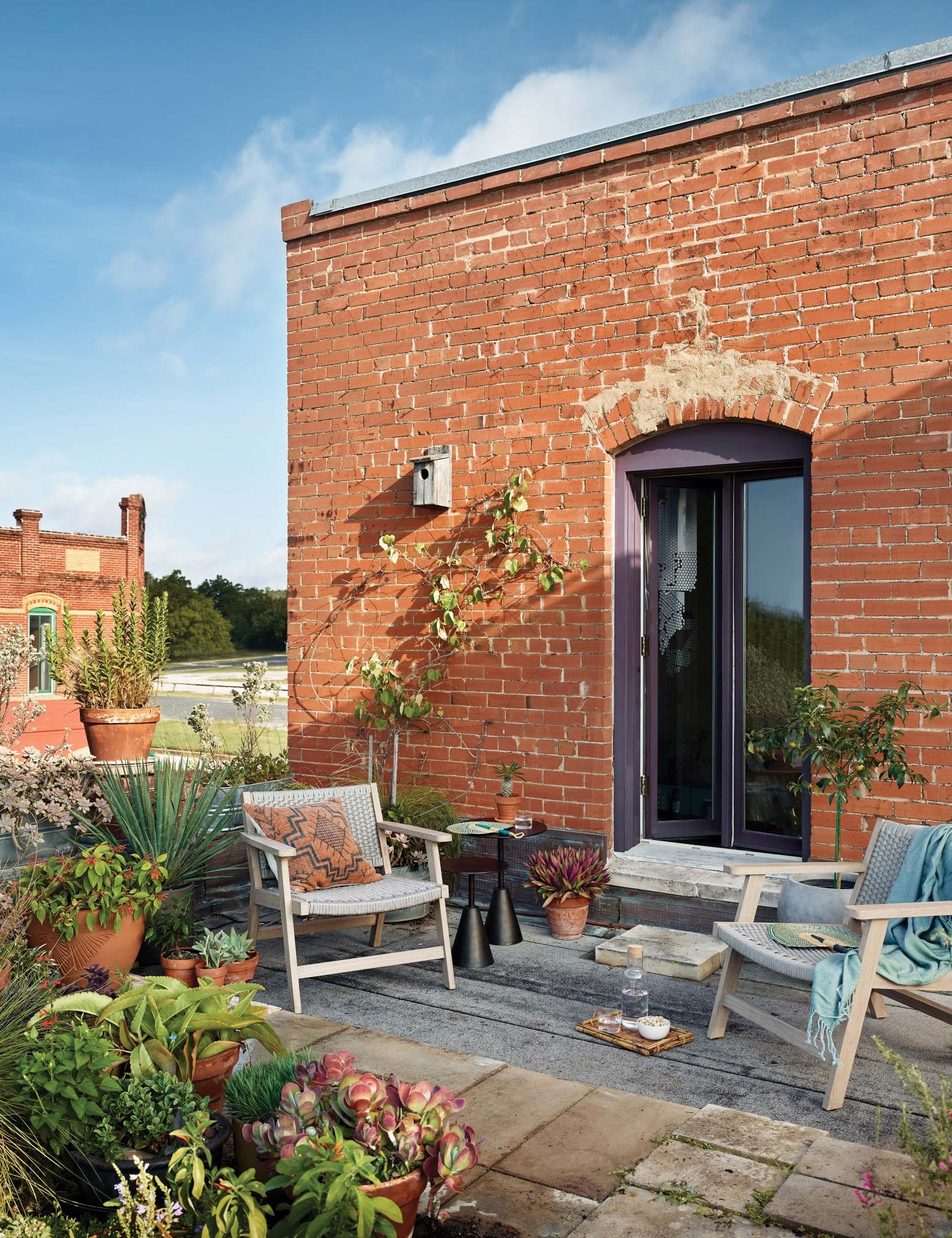 | Two Ylva Natural Indoor / Outdoor Accent Chairs sit on a rooftop outdoor space with a brick wall and various plants