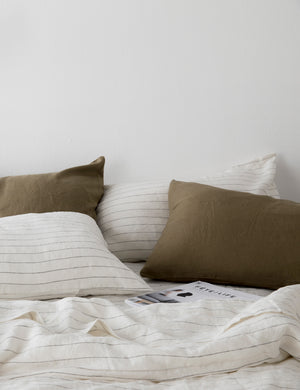 The Set of two european flax linen pencil stripe pillowcases by cultiver lay on a bed with other pencil stripe and olive green linens by cultiver