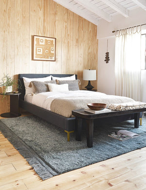  The Deva navy linen bed lays in a bedroom with a wooden paneled wall atop a gray fringed rug