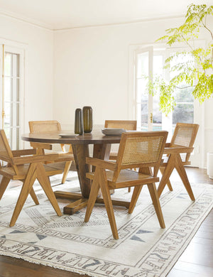 The Zehra hand-knotted ivory, gray and purple medallion wool-blend area rug achnors an airy dining room with a round wooden table with five wood and rattan arm chairs.