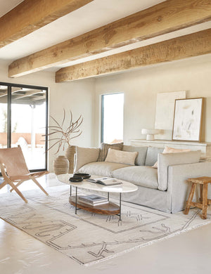 The Thomas Bina oval coffee table with white laquered top, oak shelf and steel frame in a neutral living room with a light gray slipcovered sofa and light tan leather slingback chair under wooden ceiling beams.