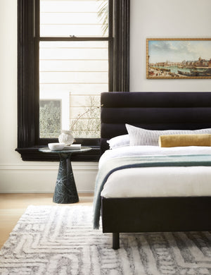 The Peregrine black and white marled striped lumbar pillow lays in a bedroom with a black marble nightstand, a black velvet framed bed, and a landscape painting