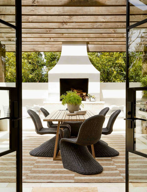 The Manila wicker weave black indoor and outdoor dining chair sits in an outdoor dining space underneath a wood paneled covering with a large white fireplace in the background.
