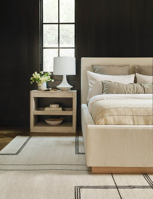 The Coulwood ivory table lamp with sculptural base sits in a bedroom atop a whitewashed wooden nightstand with a natural linen bed and patterned rug