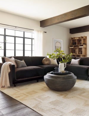 The Monroe warm gray velvet round pillow sits on a brown sectional in a living room with a round coffee table and a patterned rug