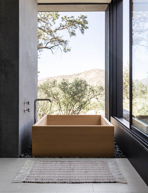 The Saskia handwoven high contrast black-and-white patterned mat with thick fringe lays on a gray tile floor in a bathroom with a square wooden bathtub and floor to ceiling windows