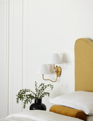 The Magdalene brass double sconce is mounted to the right of a golden linen bed on a white wall