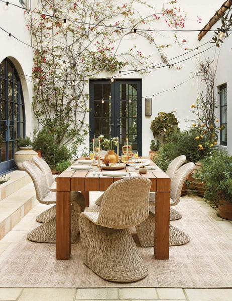 #color::vintage-white| The Manila wicker weave beige indoor and outdoor dining chair sits in an outdoor dining space surrounding a fully set wooden dining table all atop a natural patterned rug