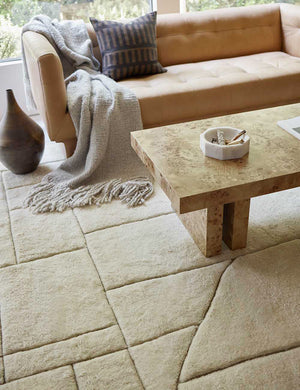 The Maleena cream hand-knotted wool and cotton area rug with abstract line tufting sits under a burl wood coffee table and a camel-colored leather sofa.