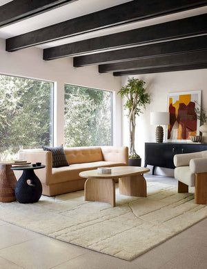 The Maleena cream hand-knotted wool and cotton area rug with abstract line tufting sits under a light wood oval coffee table, light tan leather sofa, and curved boucle accent chair.