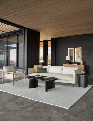 Natural Alaya Sofa sits in a living room with black walls, a black oval coffee table, a textured cream rug, and wooden ceilings