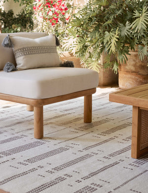 Marchesa natural and agate gray indoor and outdoor lumbar pillow with tasseled corners sits on a sofa in an outdoor space with a natural and brown patterned rug