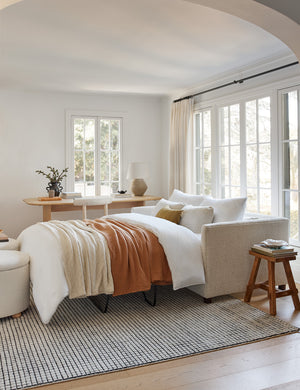 The Lotte White Basketweave queen-sized sleeper sofa with the bed pulled out sits atop a grid rug with two boucle ottomans
