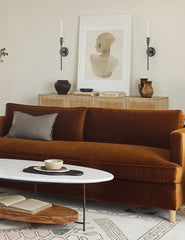 | Two Kama sleek, industrial black sconces with an aged bronze finish sits in a living room with a portrait painting in between them, a cognac velvet sofa, and a woven sideboard